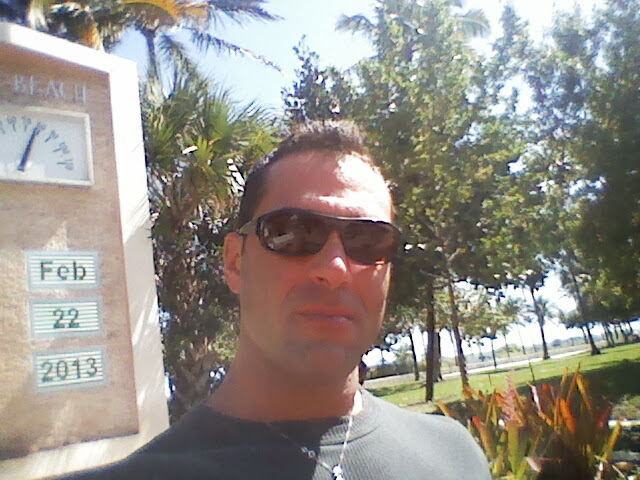 Israel Joffe taking a selfie beside a stone date-and-temperature sign on a sunny beach day