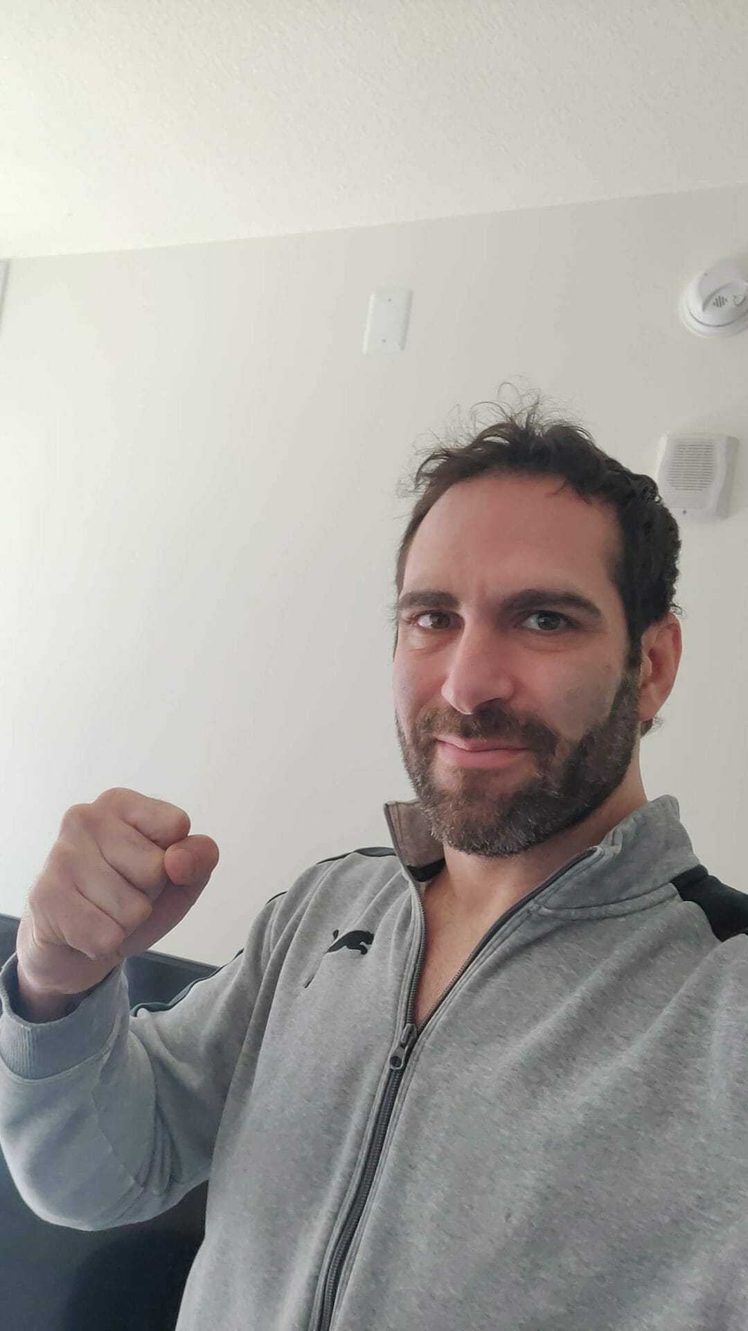 Israel Joffe in a grey zip-up hoodie with one fist raised, photographed indoors against a plain wall