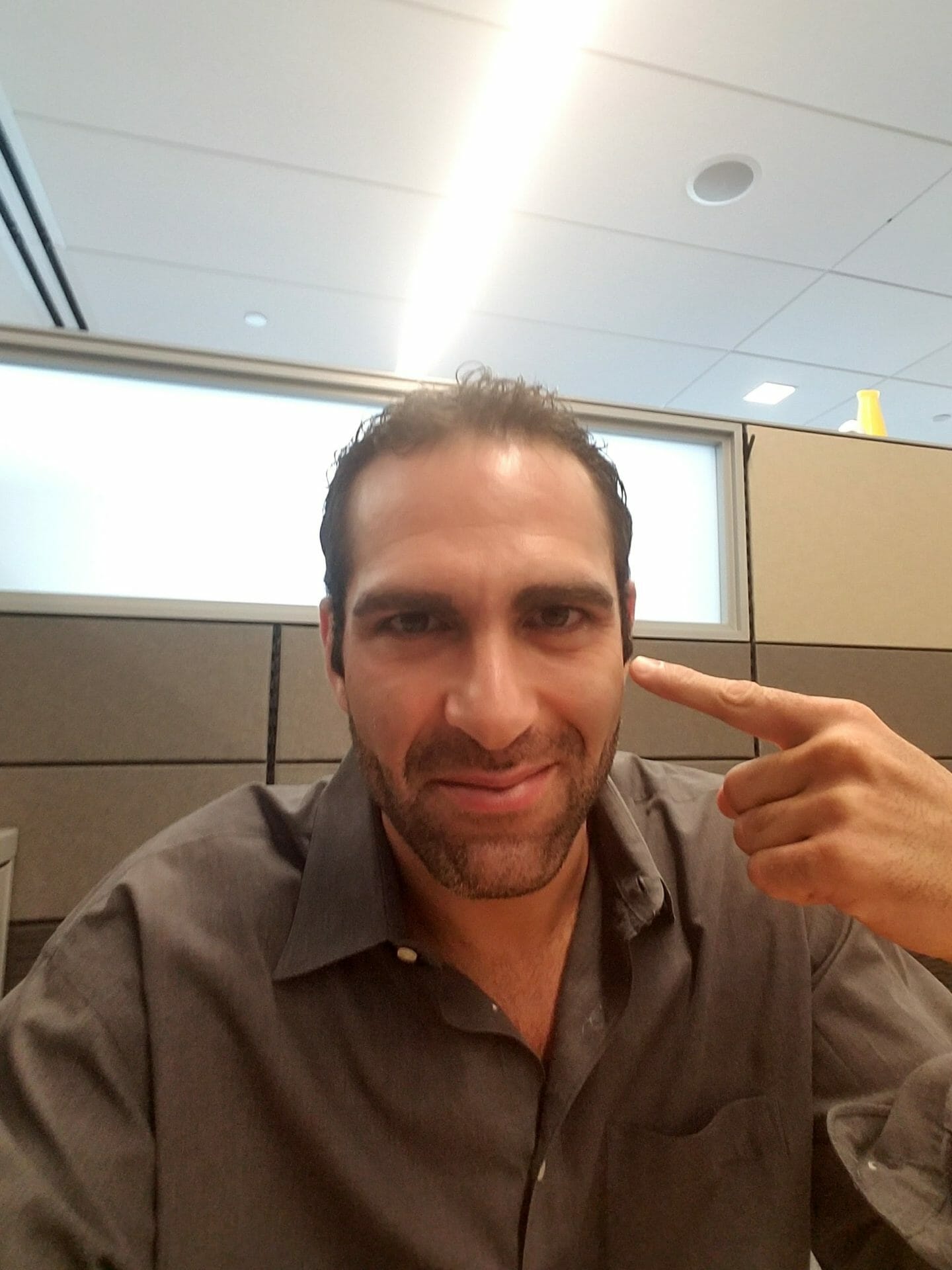 Israel Joffe seated at his desk in an office, pointing a finger at his own temple and looking at the camera