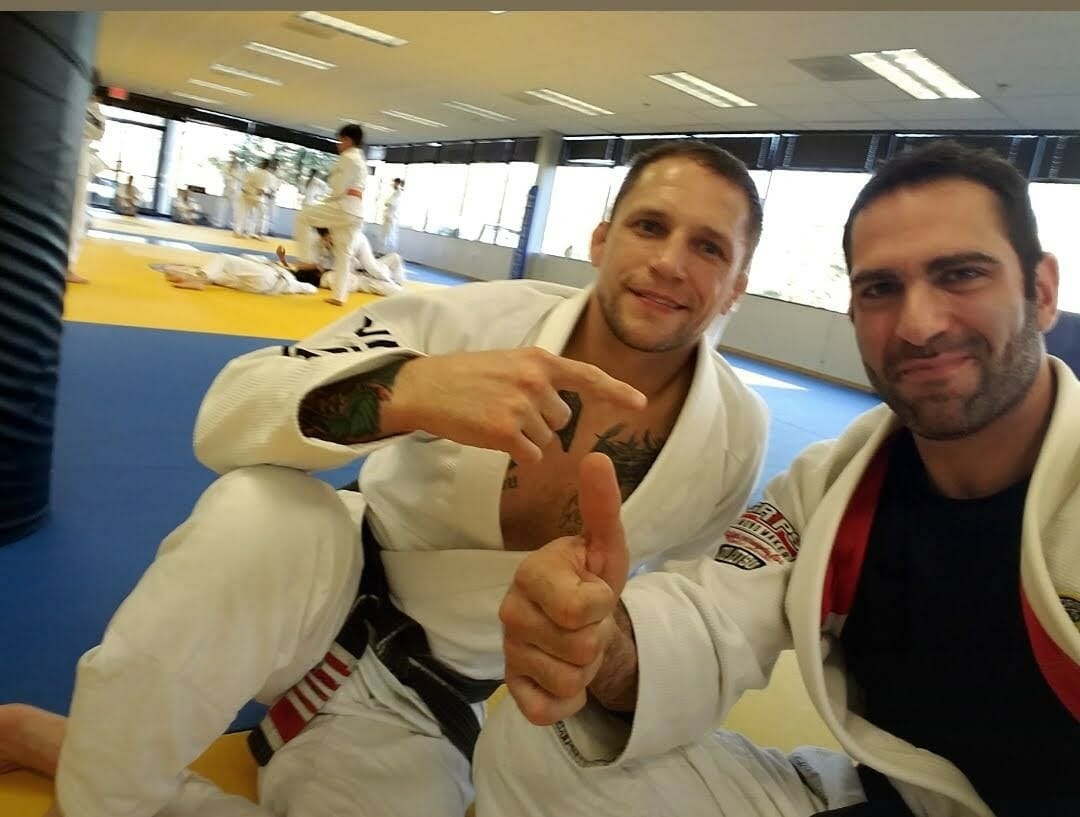 Israel Joffe in a white jiu-jitsu gi sitting on the mats with a training partner, both giving a thumbs up